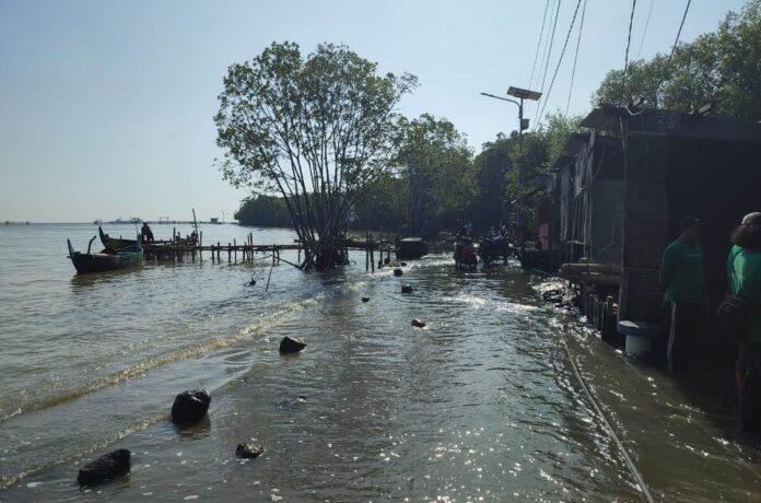 Genangan air laut mencapai kawasan warung-warung di tepi pantai, sementara perahu nelayan masih bersandar di dermaga kayu.